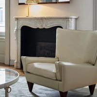 Cream leather armchair with a sleek design and walnut legs, set against a neutral backdrop and elegant marble fireplace.
