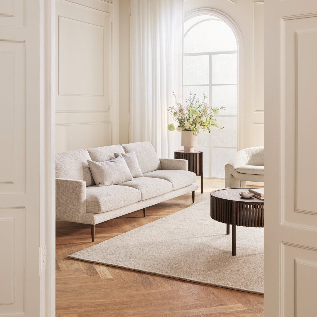 Light beige sofa with accent pillows, paired with a textured round coffee table on a soft cream rug in a bright, airy room.