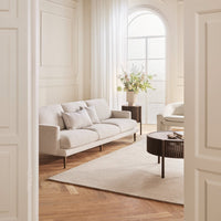 Light beige sofa with accent pillows, paired with a textured round coffee table on a soft cream rug in a bright, airy room.