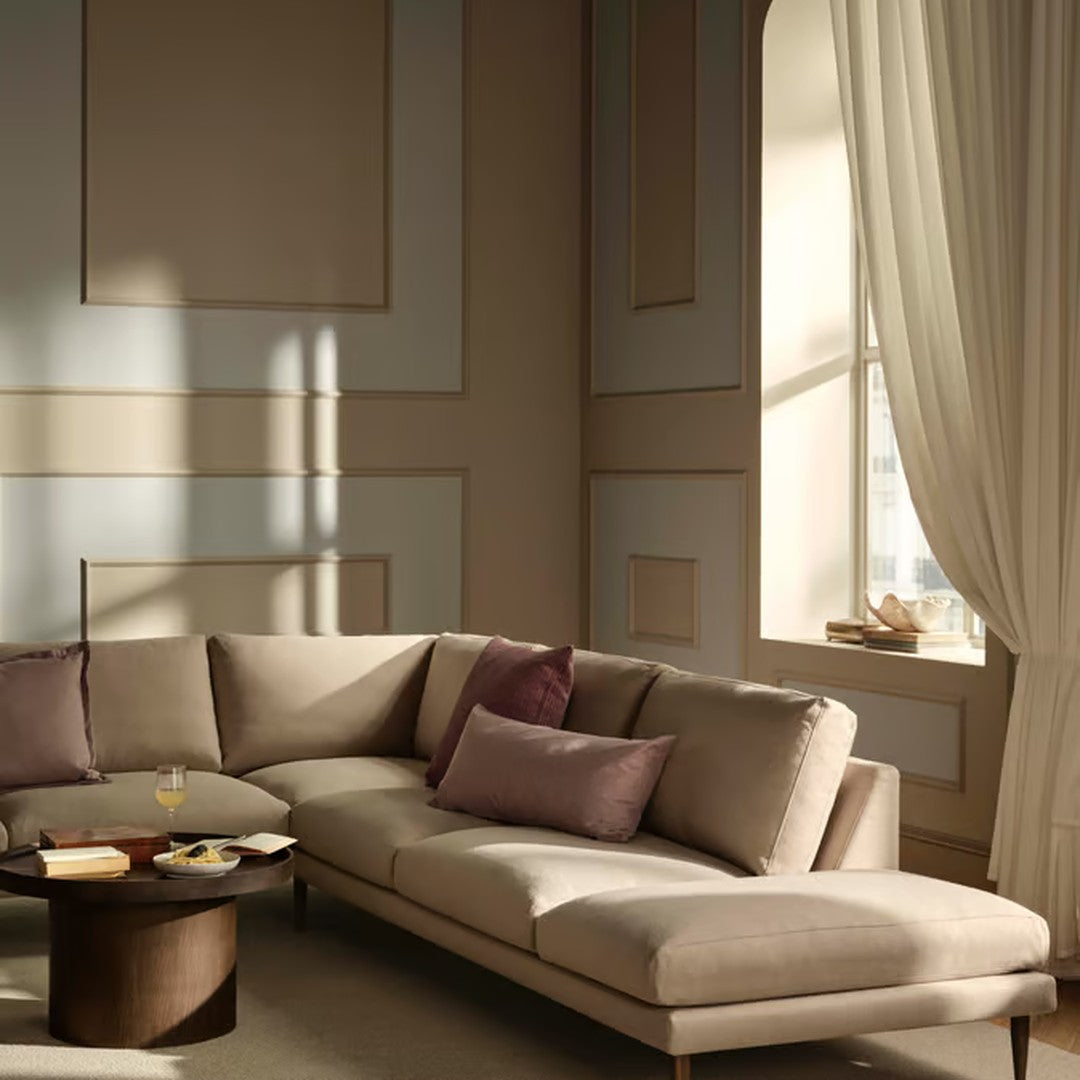 Modern beige sectional sofa with plum accent pillows, set against softly patterned cream walls and sheer beige curtains.