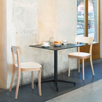 Two minimalist wooden chairs with a light natural finish around a sleek black table in a bright, airy setting.