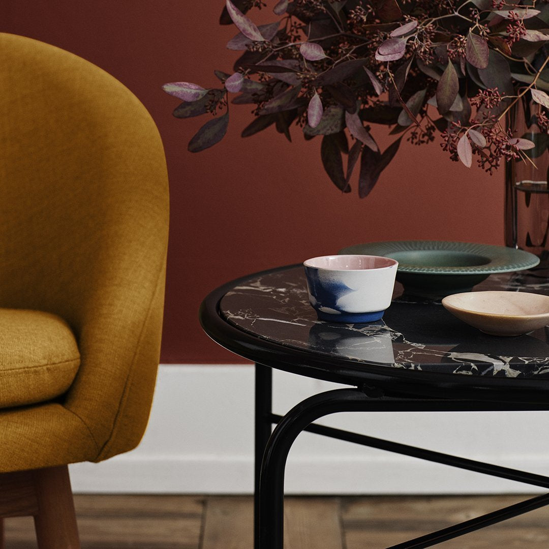 Mid-century modern chair in mustard yellow, marble table with black and white patterns, soft pastel dishware, and leafy decor.