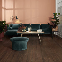 Elegant teal velvet sofa with matching round ottoman against a warm peach wall, complemented by lush green plants.