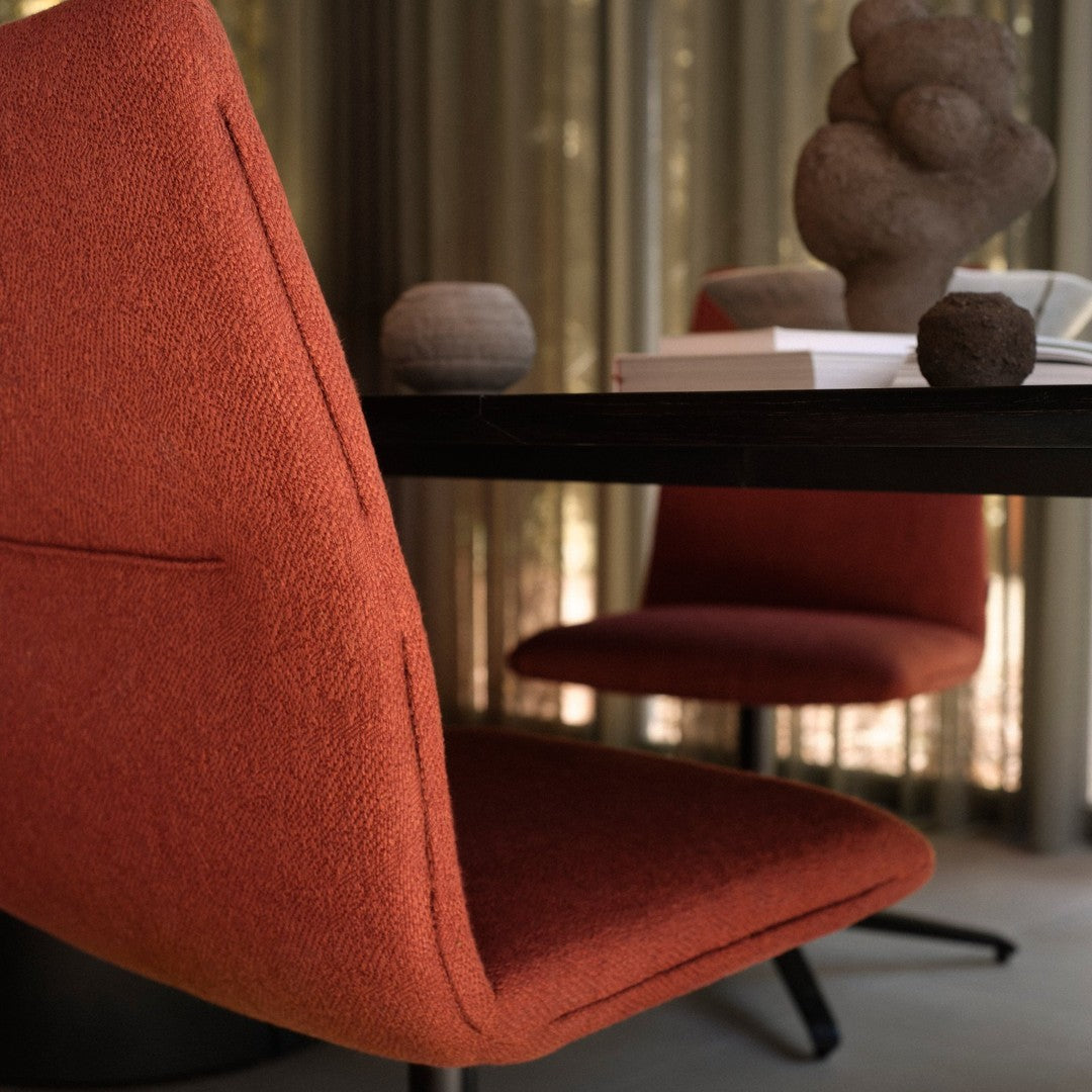 Rust-colored upholstered chair with a textured finish, set against a neutral-toned table and soft fabric backdrop.